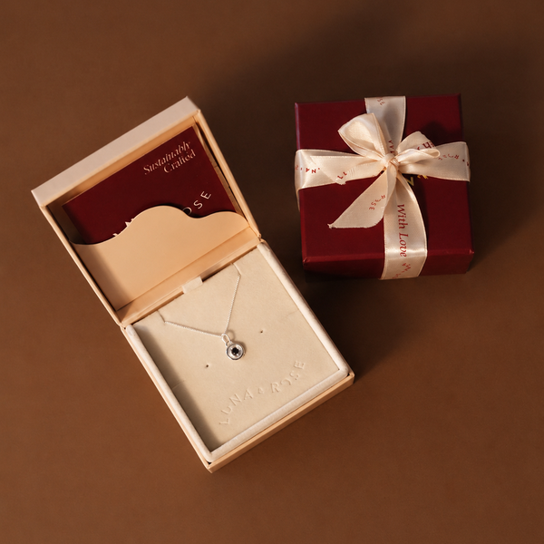 A delicate silver necklace with a small round black stone pendant is displayed inside an open cream jewelry box next to a dark red gift box tied with a cream ribbon, set against a solid brown background.