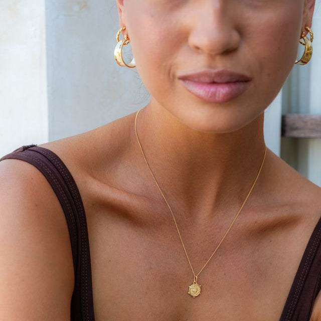 A close-up of a woman wearing a delicate gold chain necklace, paired with matching gold hoop earrings and a dark brown scoop-neck top.