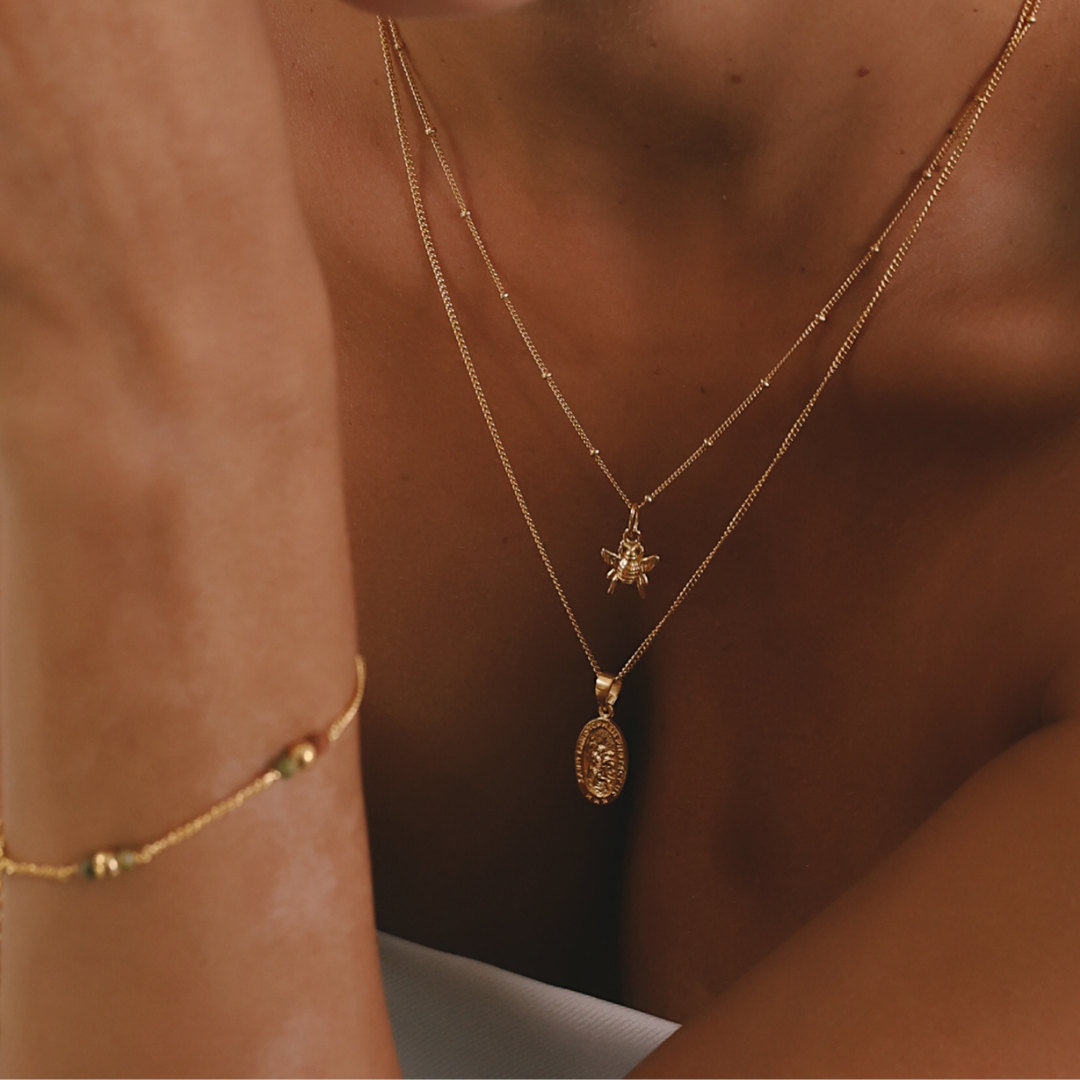 A close-up lifestyle shot of a model wearing a layered gold necklace set with a spider pendant and an oval medallion pendant, along with a matching gold beaded bracelet on her wrist.