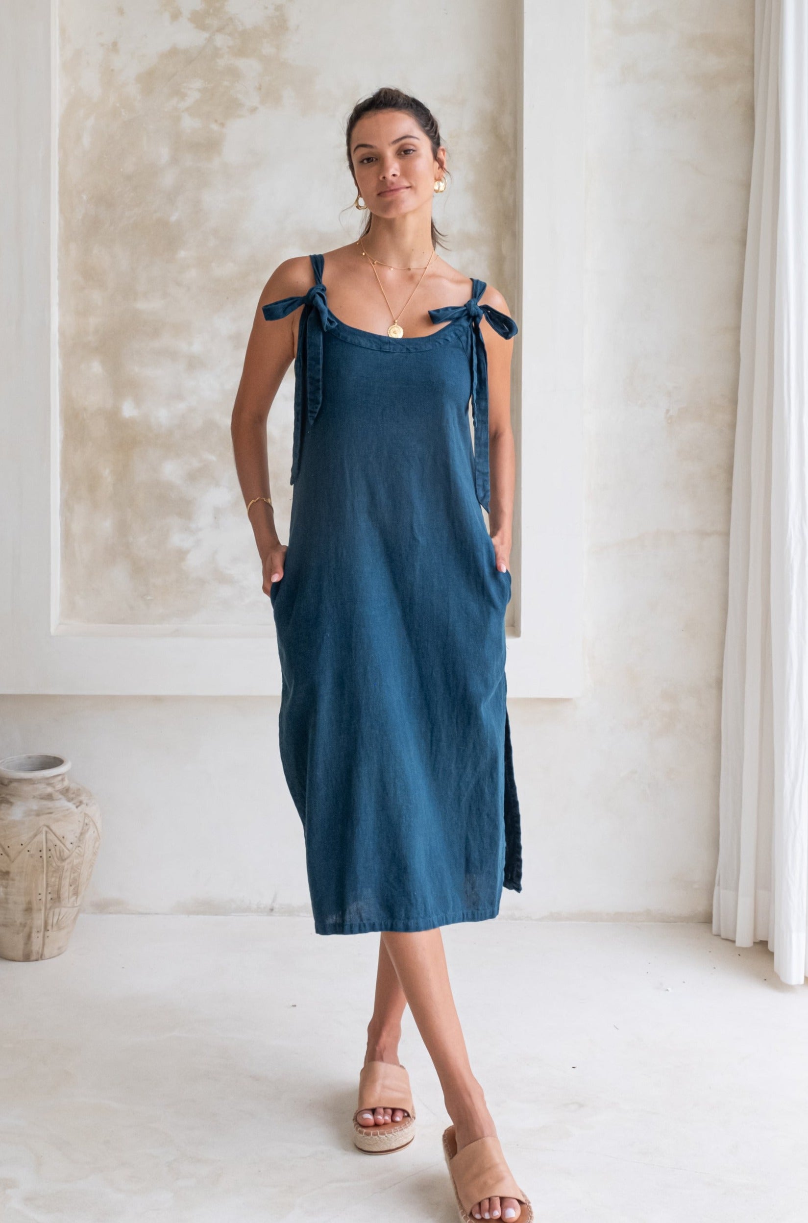 A woman in a dark blue midi dress with tied straps and beige espadrille sandals stands in a minimalist room, looking at the camera.