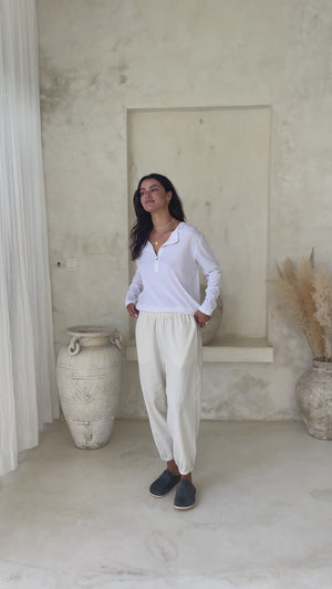 Video of a model wearing a white long-sleeve button-up top and light-colored loose pants with black slip-on shoes, accessorized with gold jewelry.