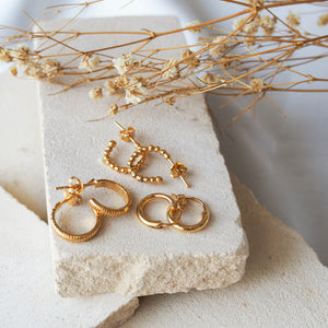 Multiple gold earrings, beaded and smooth displayed on a textured beige stone with dried flowers.