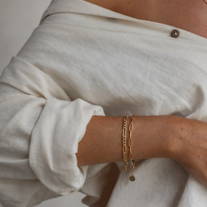 A person wearing two gold link chain bracelets stacked on their wrist, with a rolled-up sleeve.