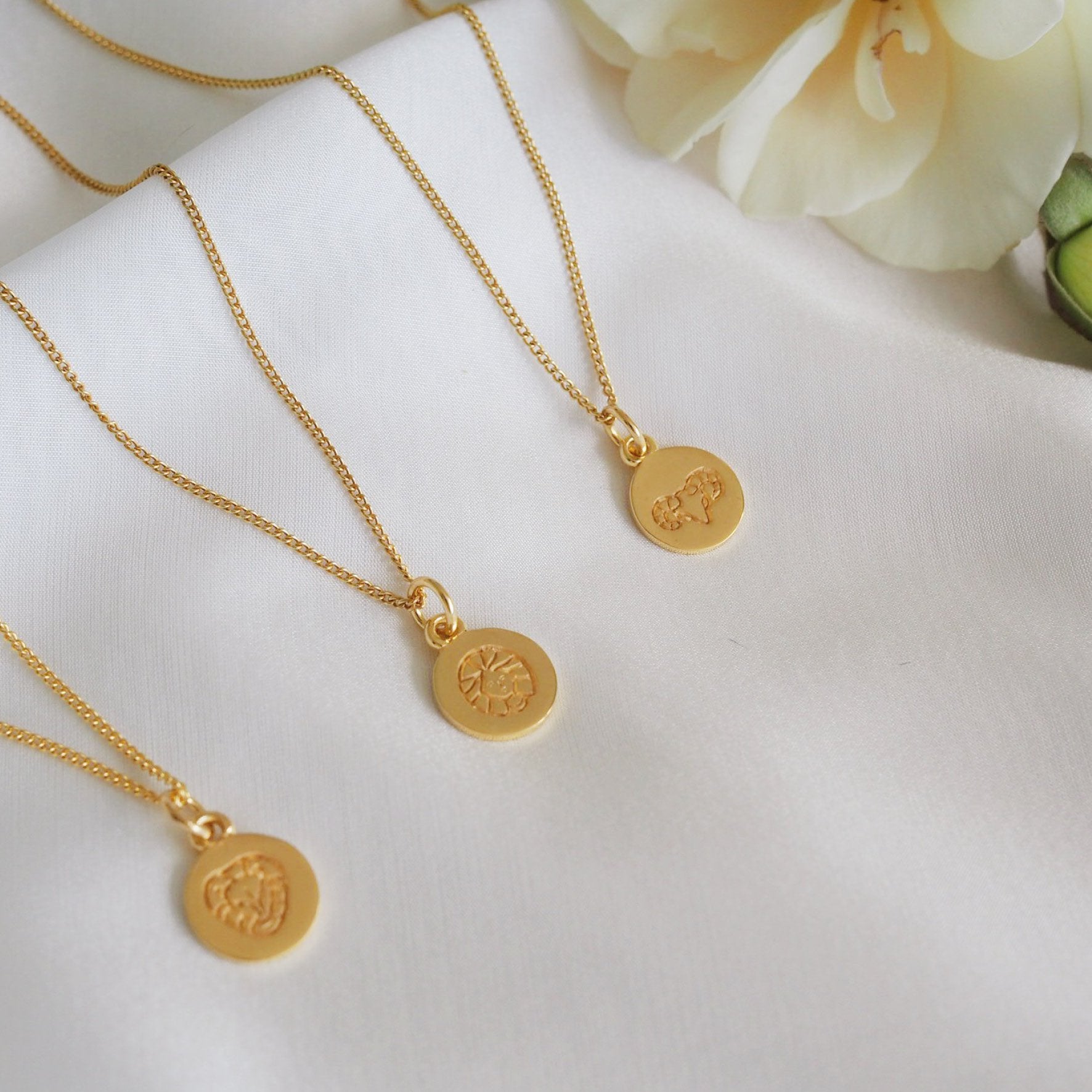 Multiple gold necklaces featuring a round pendant with a minimalist geometric design, displayed on a white fabric backdrop.