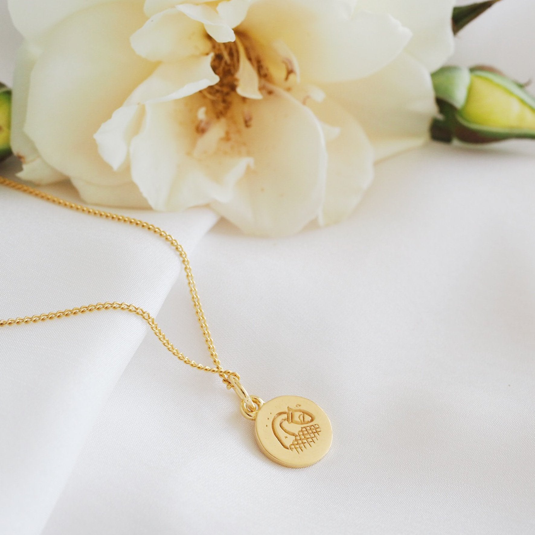 A simple gold necklace featuring a round pendant with a minimalist geometric design, displayed on a white fabric backdrop and a flower.