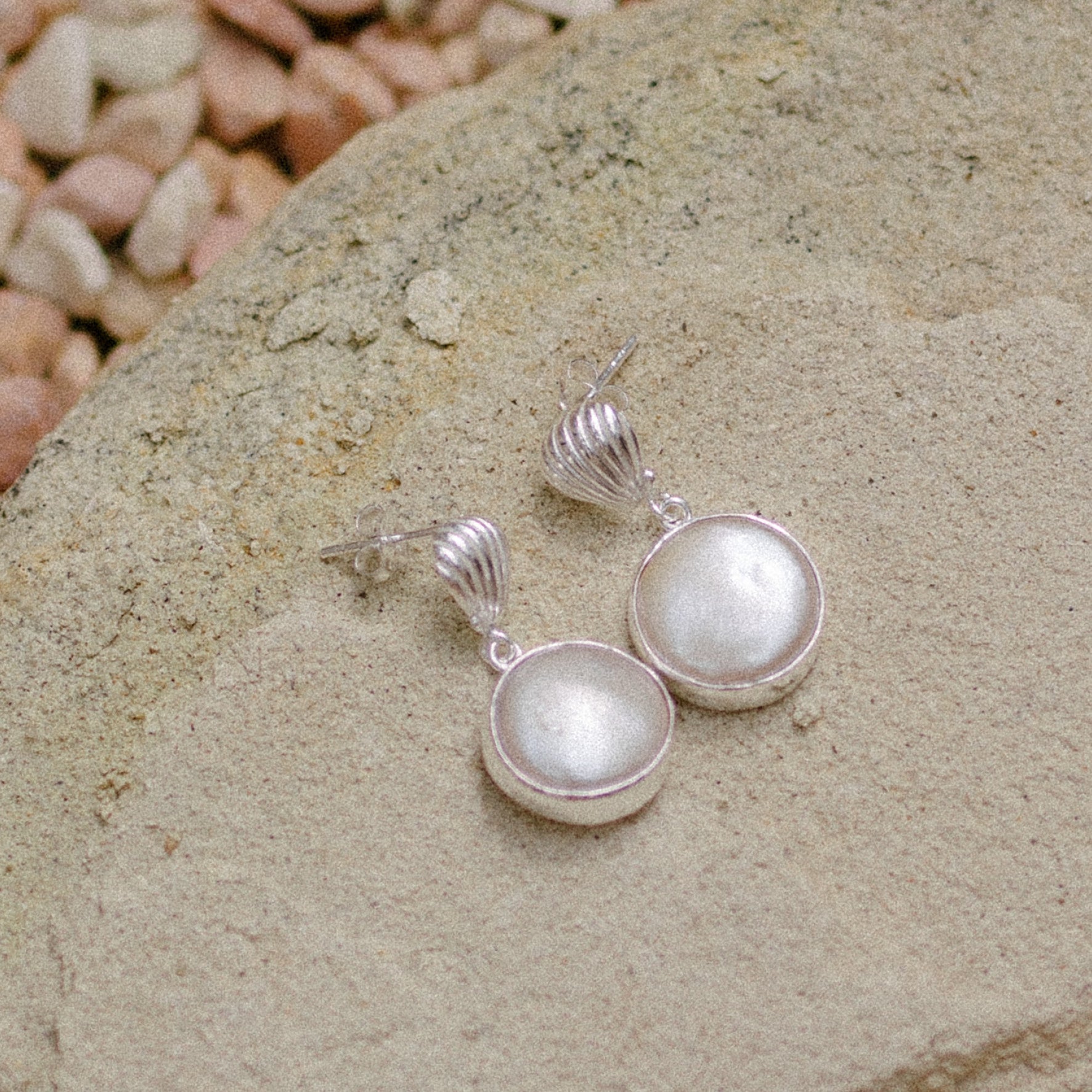 A pair of silver shell earrings with large, round white pearl pendants displayed on a stone.