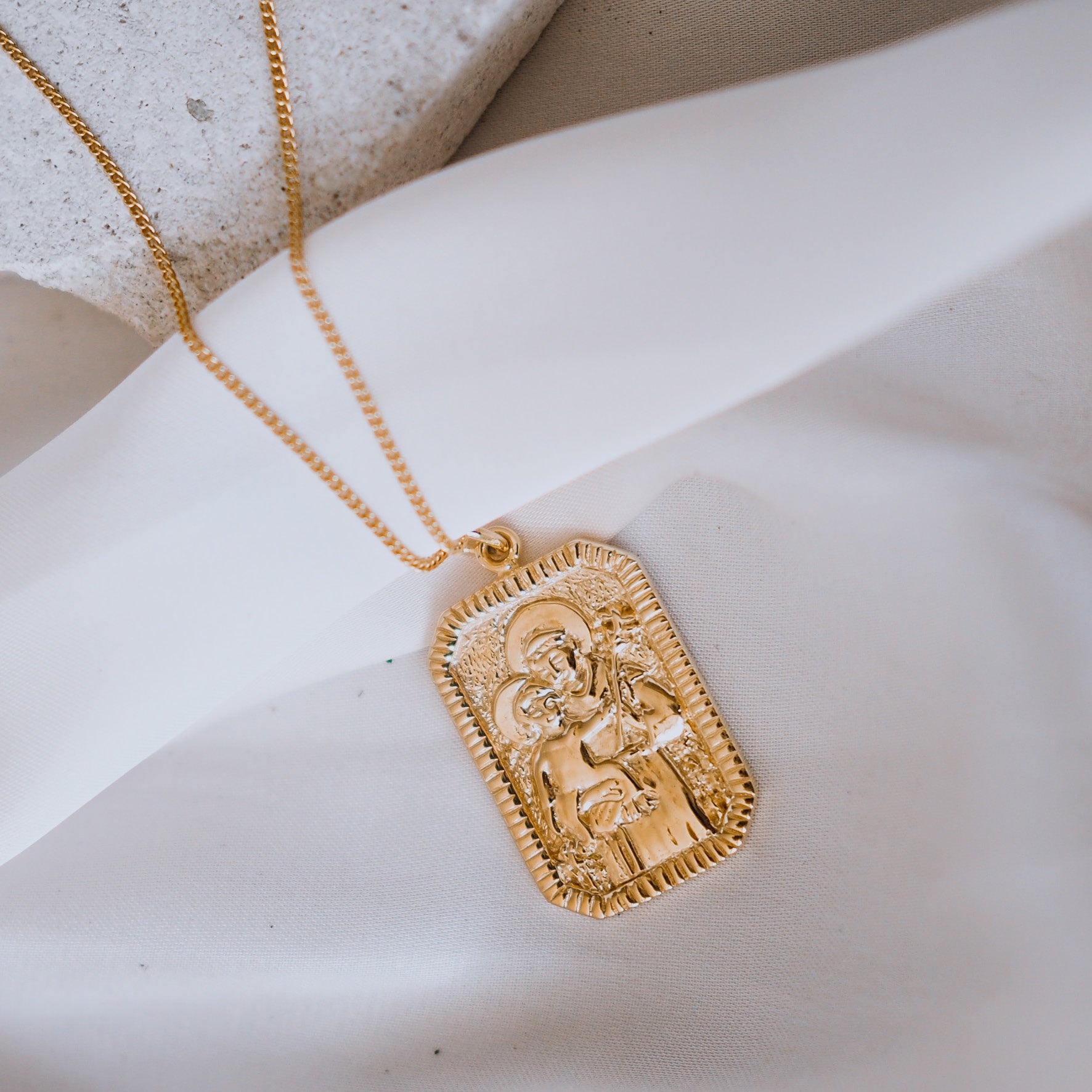 A gold necklace with a rectangular pendant featuring a raised religious figure design, displayed against a white background.