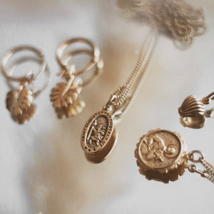 A collection of gold jewelry, including a pair of leaf-shaped hoop earrings and a necklace with an oval "Saint Christopher Protect Us" pendant, on a reflective surface.