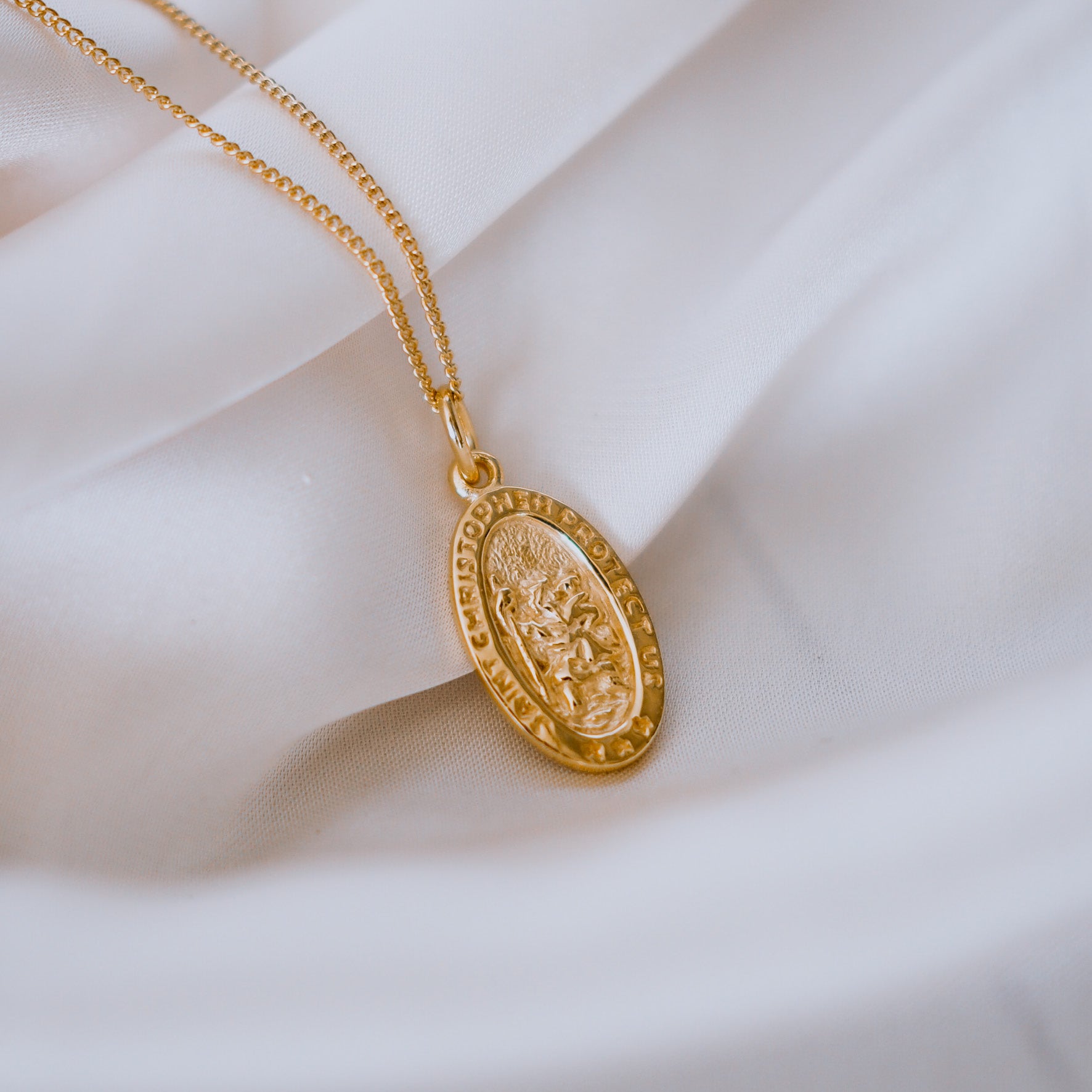 A gold necklace with an oval pendant featuring a raised religious figure design and the words "Saint Christopher Protect Us," displayed against a white satin fabric background.