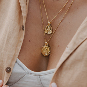 A close-up of a woman wearing layered gold necklaces with saint-themed pendants, one round with a radiant design and the other teardrop-shaped.