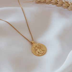 A gold pendant necklace with a detailed depiction of a saint figure, placed on a white satin fabric.