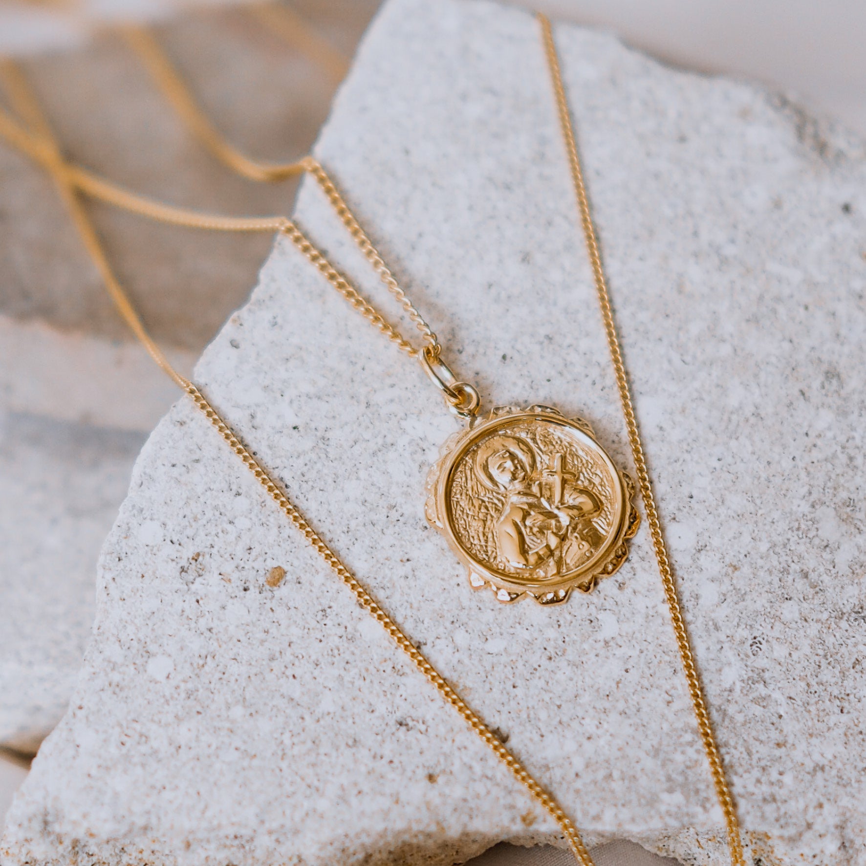 A gold necklace with a round pendant featuring a raised religious figure design, displayed against a white background.